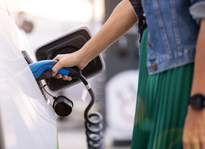A woman plugs an electric charger into the side of a white car. She is wearing a denim jacket and a green skirt. We only see from her waist down.