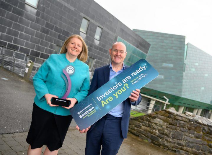 A man and a woman standing together with a brick wall in the background. The man is holding a sign about the Seedcorn competition, while the woman is holding an award trophy.