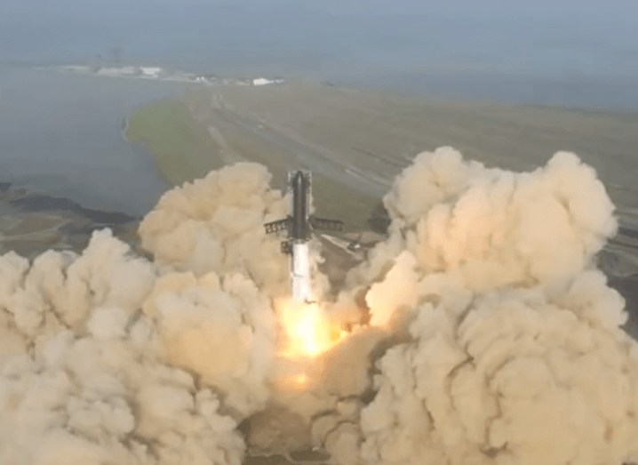 An aerial view of the Starship rocket taking off with smoke around it and a jet of flame under it.