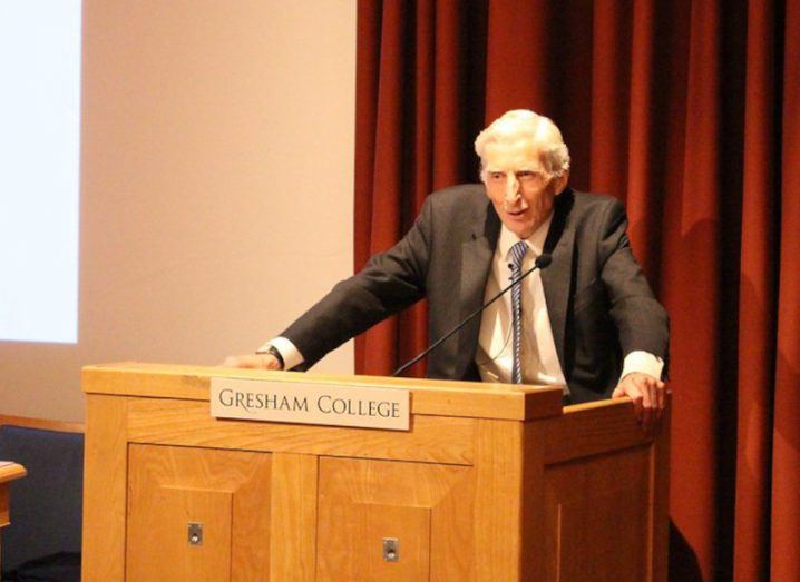 Astronomer Martin Rees standing behind a lectern speaking. There is a red curtain on stage behind him.