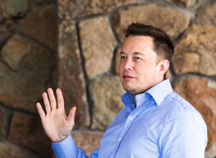 Elon Musk wearing a blue shirt, waving in front of a brown stone wall.
