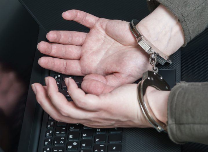A person's hands with handcuffs on the wrists, on top of a laptop keyboard. Used as a concept for cybercrime.
