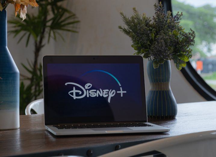 The Disney+ logo on a laptop screen, which is on a wooden table next to some house plants and a window in the background.