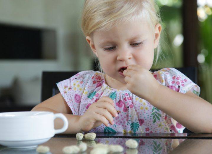 A little girl sitting at a table eating shelled peanuts. There is a white cup in front of her.