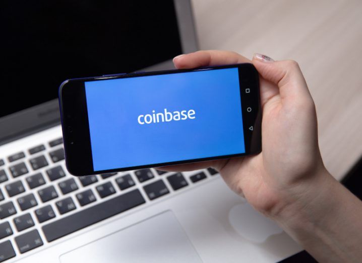 The Coinbase logo on a smartphone screen that is held in a person's hand, with a laptop and table behind it.