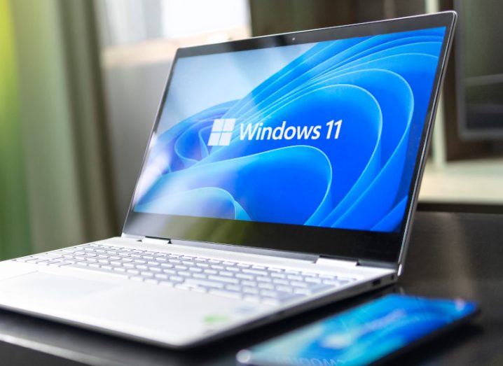 The Windows 11 logo on the front of a laptop screen. The laptop is laying on a table next to a mobile phone.