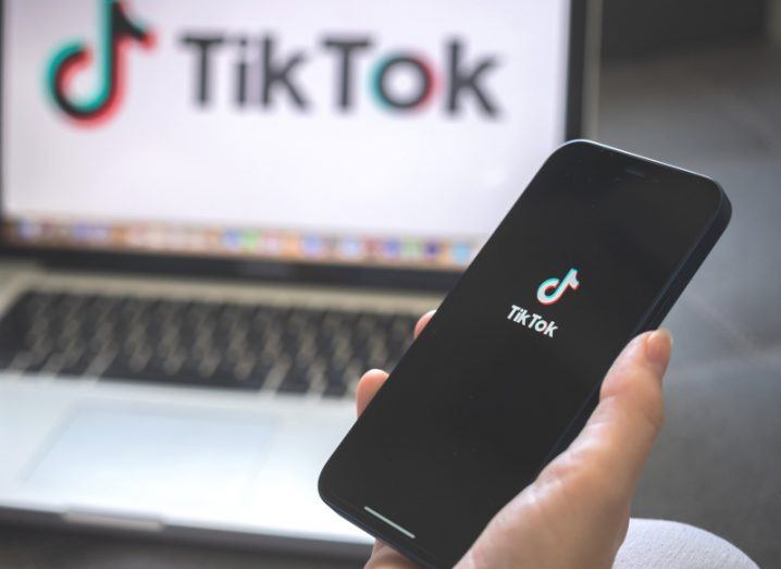 The TikTok logo on a smartphone screen, held in a person's hand. A laptop is in the background that also has the TikTok logo on the screen.