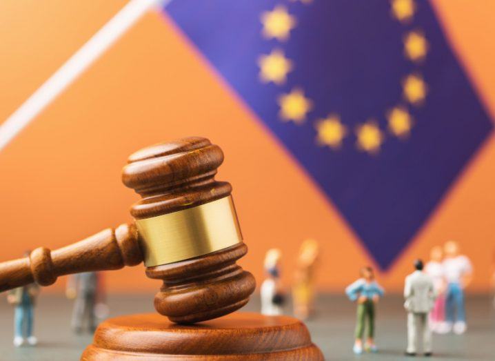 A large wooden gavel with the EU flag behind it and little toy plastic figurines like people standing around the gavel and flag.