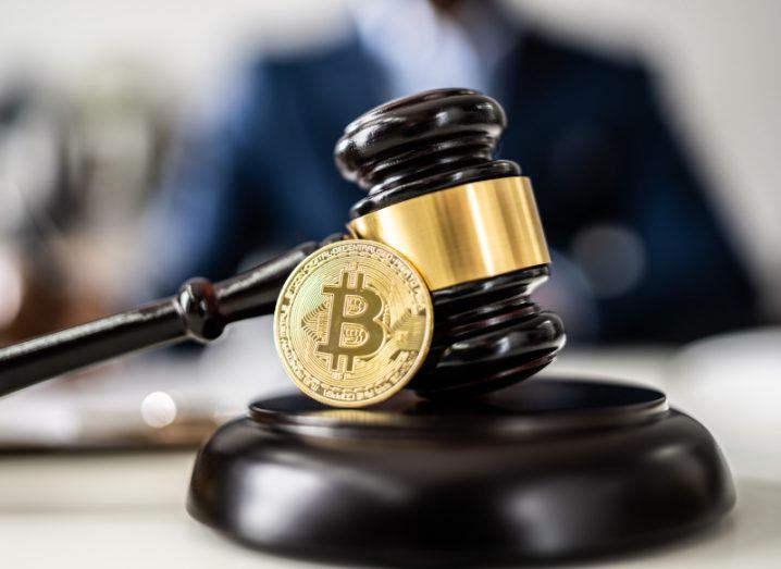 A bitcoin in front of a gavel laying on a table, with a person in a suit in the background. Used as a concept for crypto regulation.