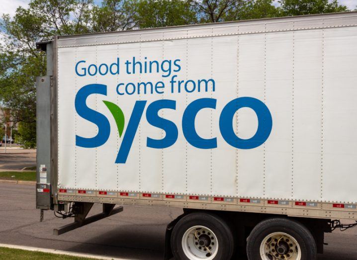 The side of a truck with the phrase "Good things come from Sysco" written on it, with trees in the background.