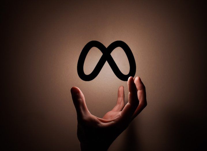 The Meta logo on a brown background with a hand reaching up to grab it.