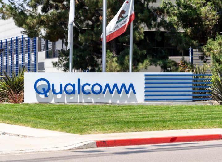 The Qualcomm logo on a sign behind a patch of grass, with a road in the front of the image. There are trees, flags and a building behind the sign.