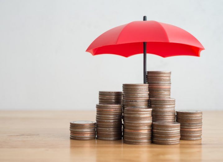 Multiple stacks of coins with a red umbrella above them, resting on a wooden table.