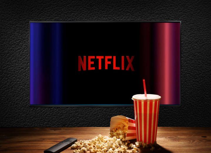 The Netflix logo on a large TV, behind a wooden table with a bag of popcorn, a drink with a straw and a remote on the table.