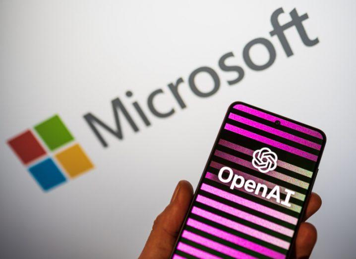 A smartphone with the OpenAI logo on it. The phone is in front of a white background that has the Microsoft logo on it.