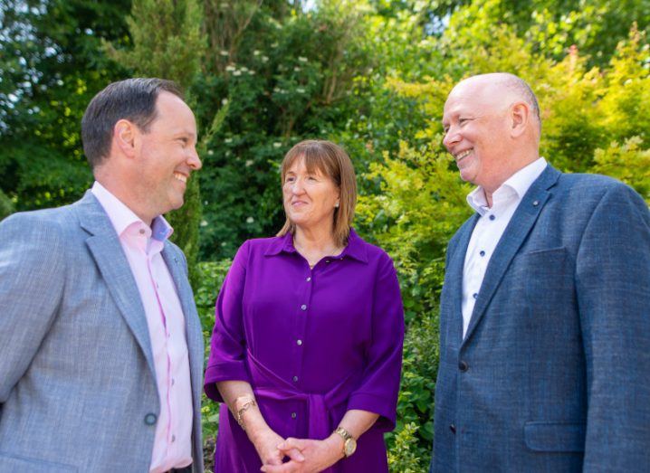 Two men and a woman standing together smiling at each other, with green trees in the background. They are executives of Milestone Solutions and PM Group.