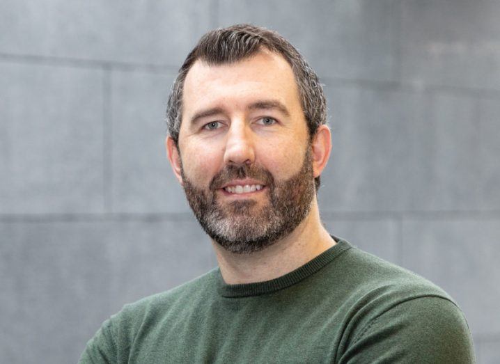 A man in a green jumper, with a grey brick wall behind him. He is Dr Andy Hogan.