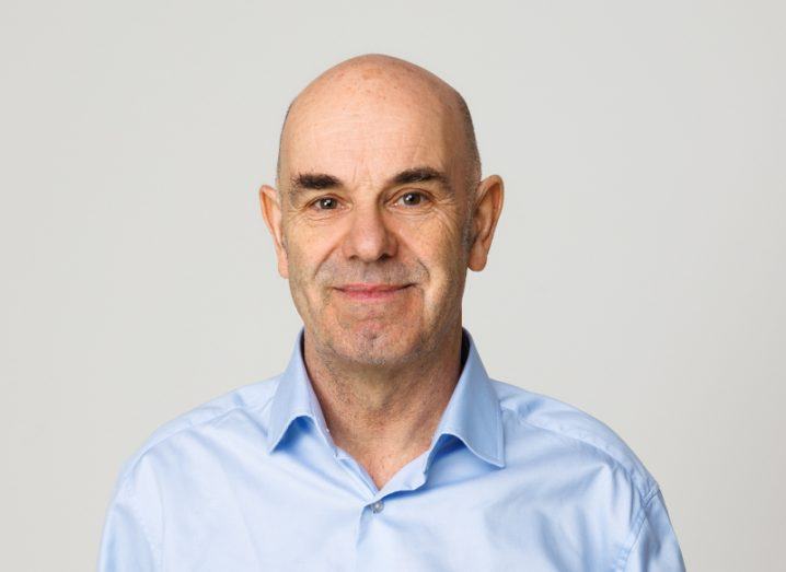 A man in a light blue shirt smiles at the camera with a light grey background.