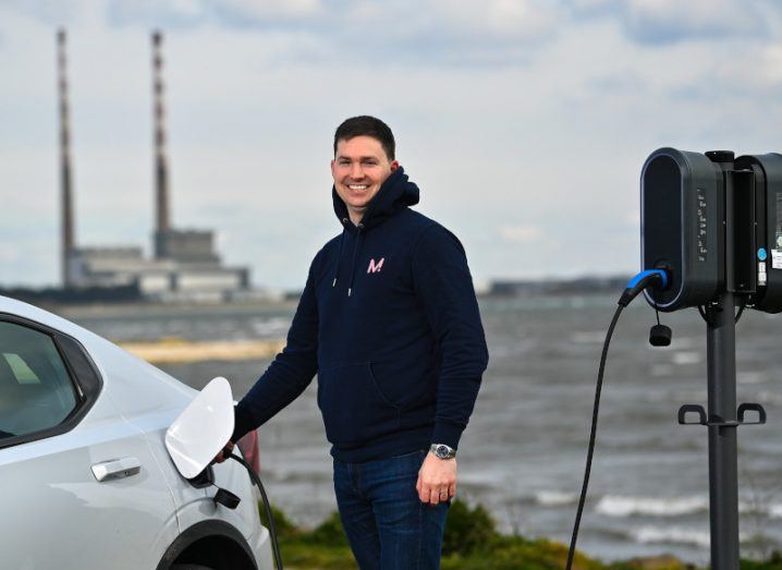 A man charging an electric vehicle at a charge point, with the sea and a power plant in the background. He is Ricky Hill of Monta.