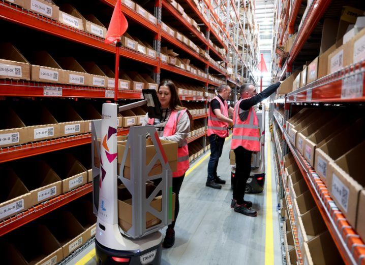 Three people working at a warehouse with a robot in front of one of the workers, holding cardboard boxes.
