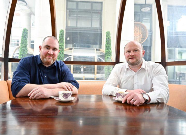 Two men sitting at a table, with windows and a building in the background. They are the co-founders of Weev.