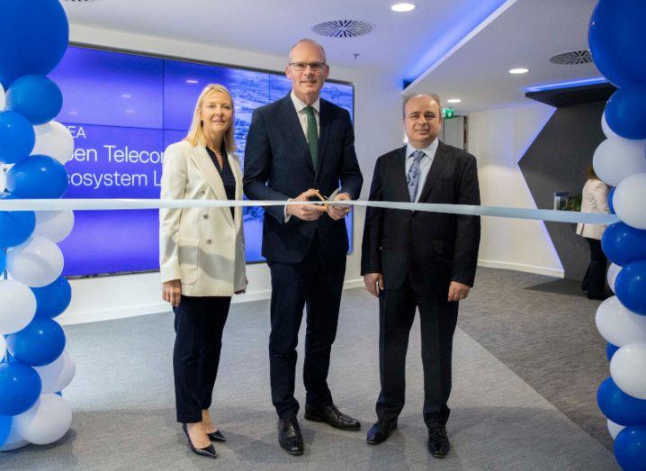 Simon Coveney, TD cuts a ribbon at Dell office. Catherine Doyle stands on his right in a cream blazer and a man in a suit stands to his left. Blue and white blues flank them.