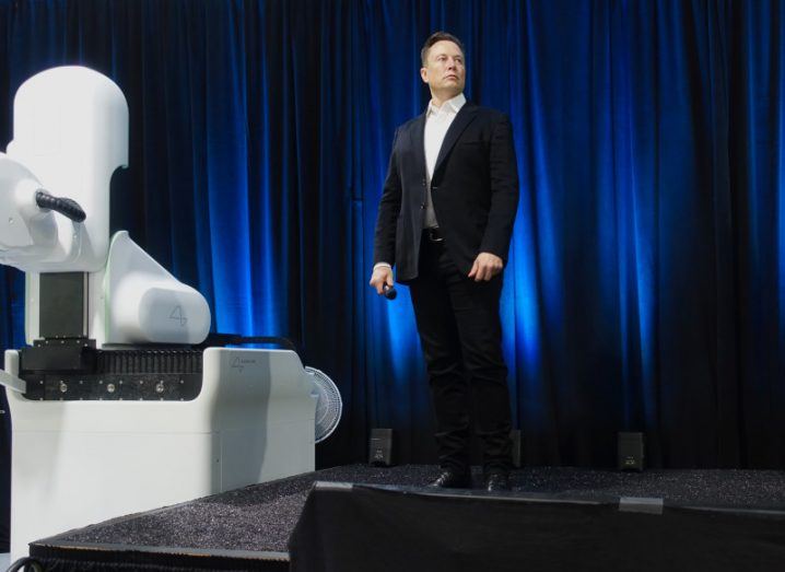 Elon Musk in a suit standing on stage with a Neuralink device in front of him.