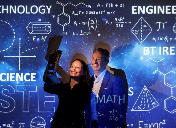 A woman holds a BT Young Scientist award in her hands and raises it up while a man stands next to her and smiles. Behind them is a wall with mathematical and scientific equations and symbols projected on it.