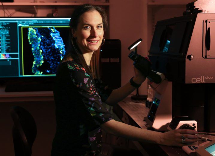 A woman smiles at the camera while standing at a large microscope in a lab. There is a microscopic image on a screen behind her.