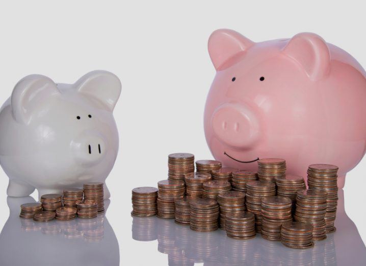 Two piggy banks with coins in front of them, in a white background. The pink piggy bank on the right has a larger amount of coins than the smaller grey piggy bank on the left.