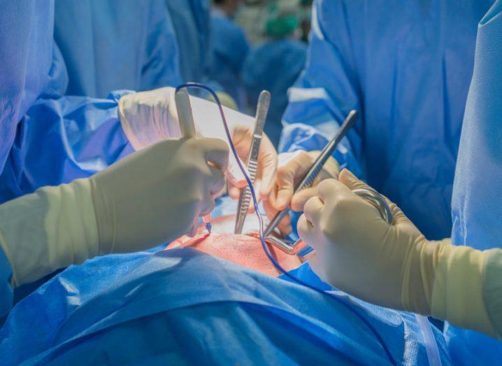 An image of surgeons performing surgery. The image is zoomed in to the hands with scalpels with the patient's skin and blue scrubs visible.