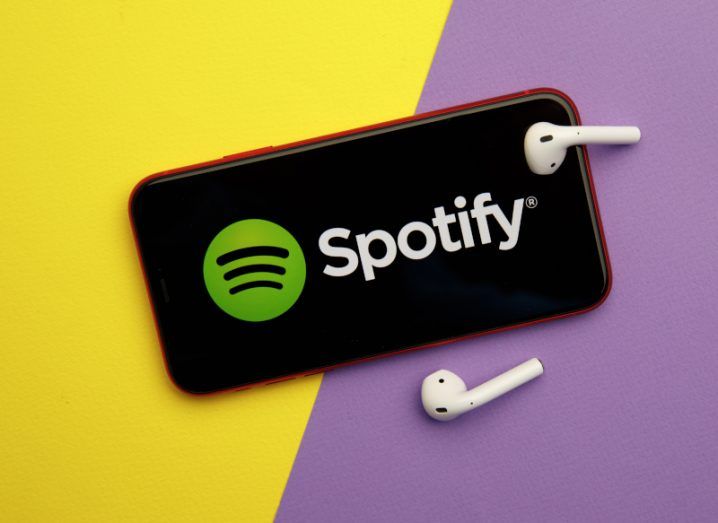A phone with Spotify open on it and wireless earphones beside it. The background is purple and yellow.