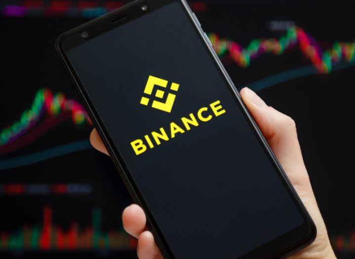 The Binance logo on a smartphone screen, held in a person's hand with another screen in the background showing stock prices.