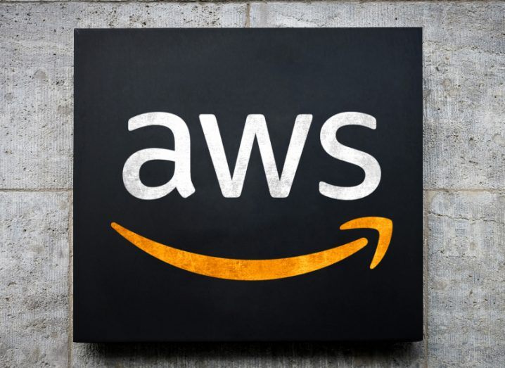 The Amazon Web Services AWS logo in white letters on a black square. The logo sign is hanging on a grey wall.