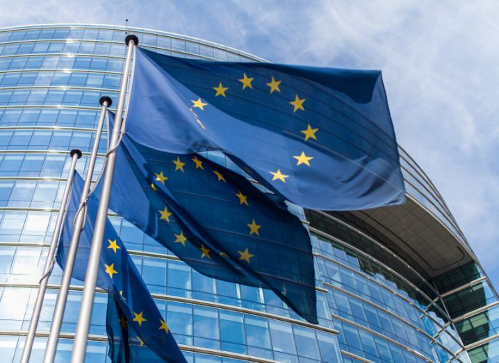 Three EU flags waving in front of a large windowed building, with the sky visible in the background.