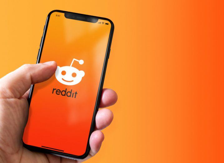 A smartphone with the Reddit logo on the front, held in a person's hand and an orange background behind it.