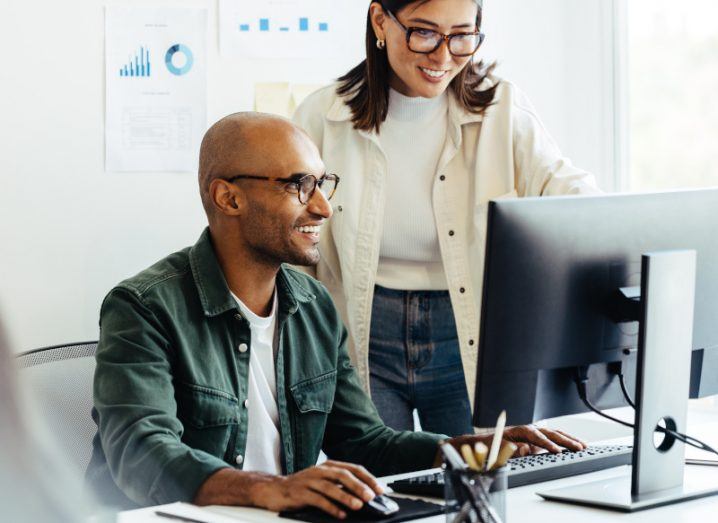 Man sitting down at a computer while a woman stands beside him helping, symbolising the benefits of automation.