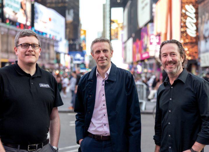 Three men stand together smiling at the camera with what appears to be Times Square in the background.