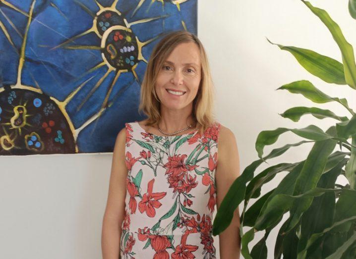 Prof Paula Petrone standing in front of a white wall with a painting hanging on it partially visible and a plant in the corner.
