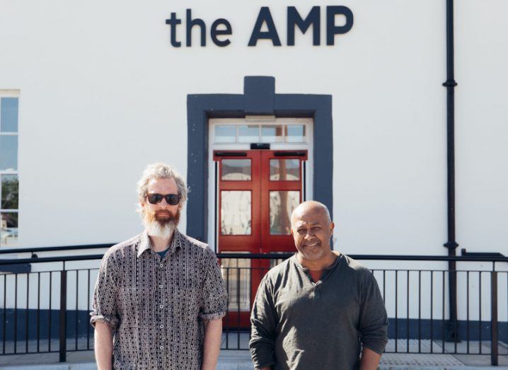 Two men standing outside the front of a white painted building with a sign reading AMP on it and railings surrounding its perimeter.
