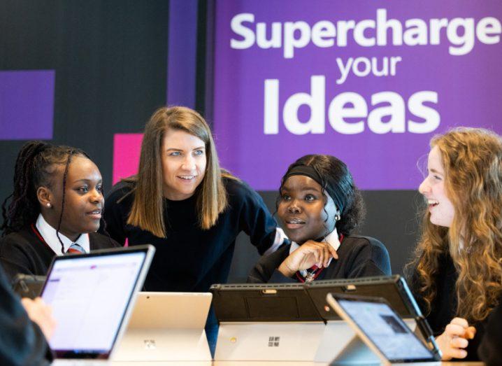 Three students speaking with a woman by a table with a wall in the background that has 'Supercharge your ideas' written on it.