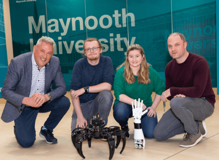 Three men and a woman crouching over a black robot, with a wall behind them that says Maynooth University.