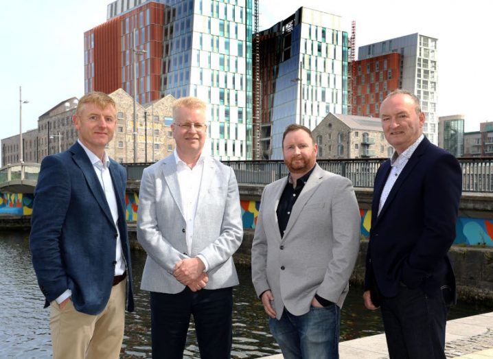 Four men standing together with a river and buildings in the background. They are part of Tekenable and Tether.