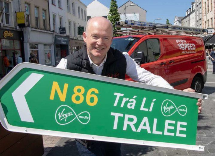 A man holding a signpost for Tralee, with a red Virgin Media van and a town in the background. He is Tony Hanway of Virgin Media.