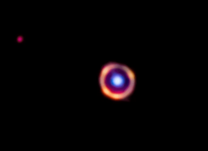Bright blue circle surrounded by orange dots forming a larger circle. Background is pitch dark with a small red dot to the top left of the image taken by the James Webb telescope.