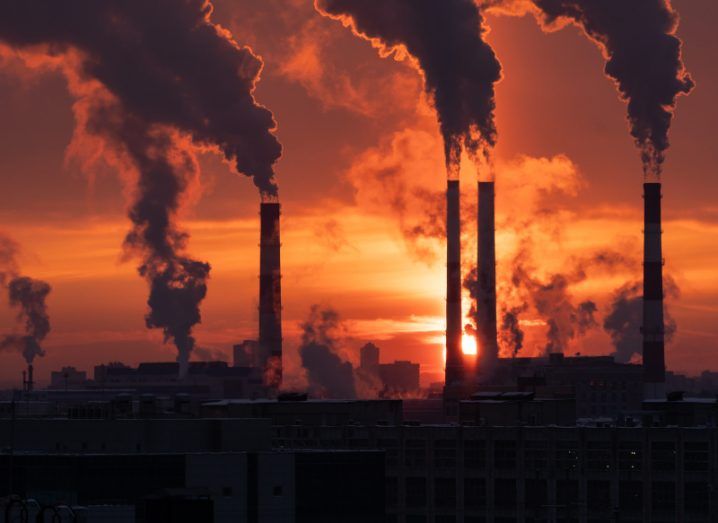Many dark chimney stacks emitting CO2 at a power plant with an orange sunset in the background.