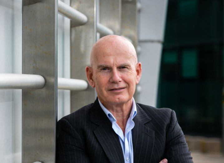 Headshot of a man in a suit with his arms crossed, standing in front of a tall grey railing. He is Ian Brown of Integrity360.