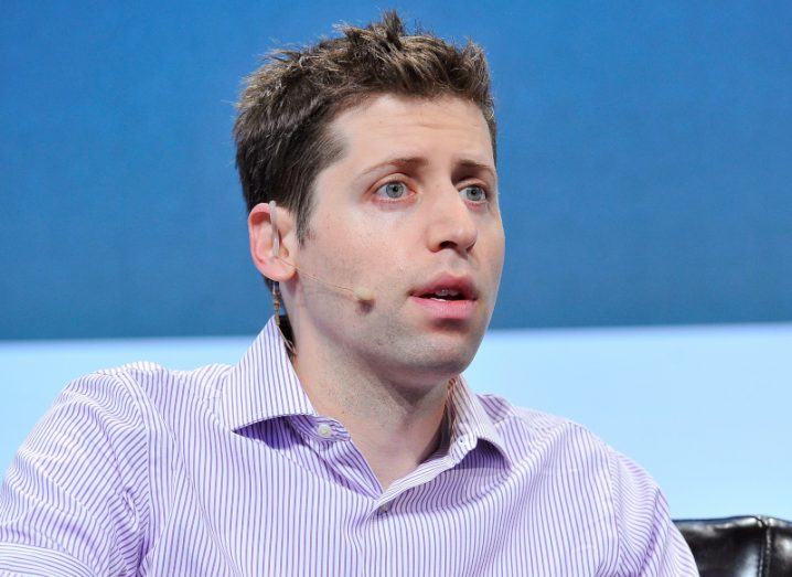 A man with a microphone connected to his ear, sitting on a stage. He is Sam Altman.
