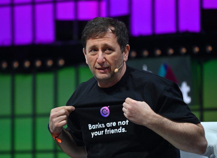 A man standing on a stage, wearing in a black t-shirt that says "banks are not your friends" with a green and purple background behind the man. He is Alex Mashinsky, the founder and former CEO of Celsius Network.