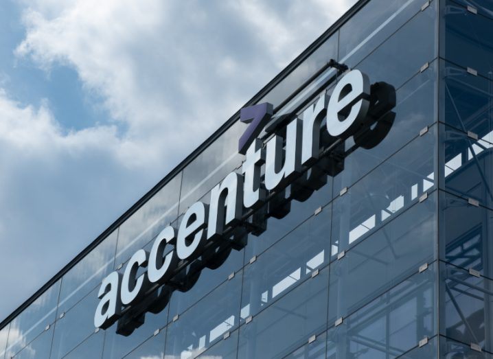 The Accenture logo on top of a glass building against a cloudy sky.